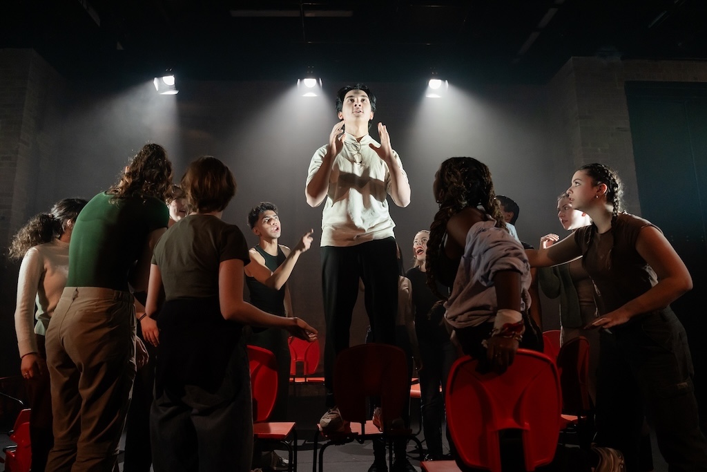 Action shot of the spring 2024 Shakespeare in Performance at RADA final presentation of Julius Caesar. An actor stands on a bright orange chair in mid-performance, lit by spotlights overhead, while other actors surround him on stage.